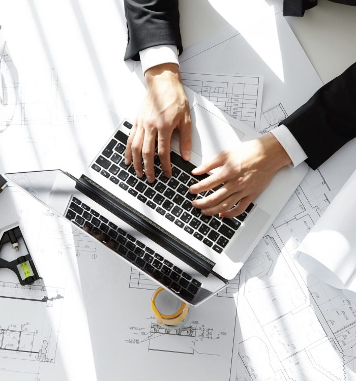 top-shot-architect-contractor-sitting-desk-with-laptop-computer-sketches-scale-model-house-blueprint-rolls-ruler-entering-data-while-working-new-housing-project-his-office-min